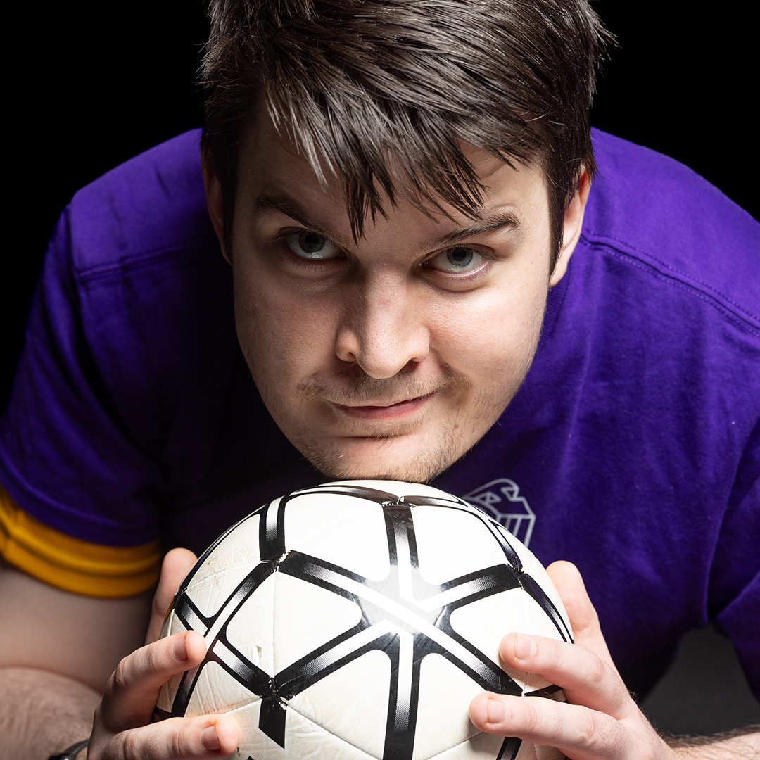 Brendan - Harrow College Level 3 Sport student - he is leaning in and holding a football. He is smiling and wearing a purple and yellow top.