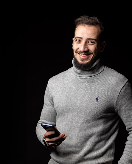 Ahmad - an IT Harrow College student - he is wearing a grey polo neck from Ralph Lauren and has a goatee beard. He is smiling and holding a mobile phone.
