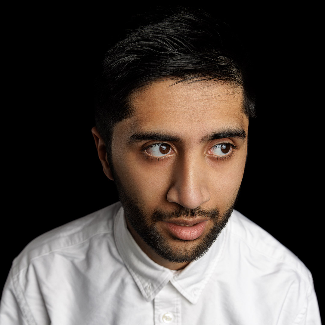 Kabir has his mouth slightly open and looking off to the side. He has a beard and dark hair and is wearing a white shirt. 