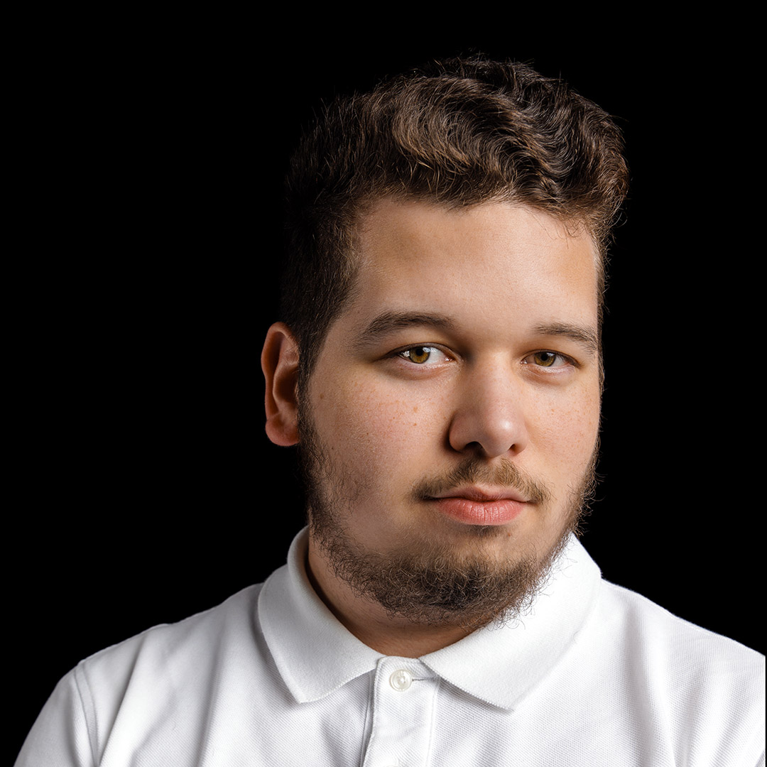 Zak has curly hair and a beard. He is wearing a white shirt.