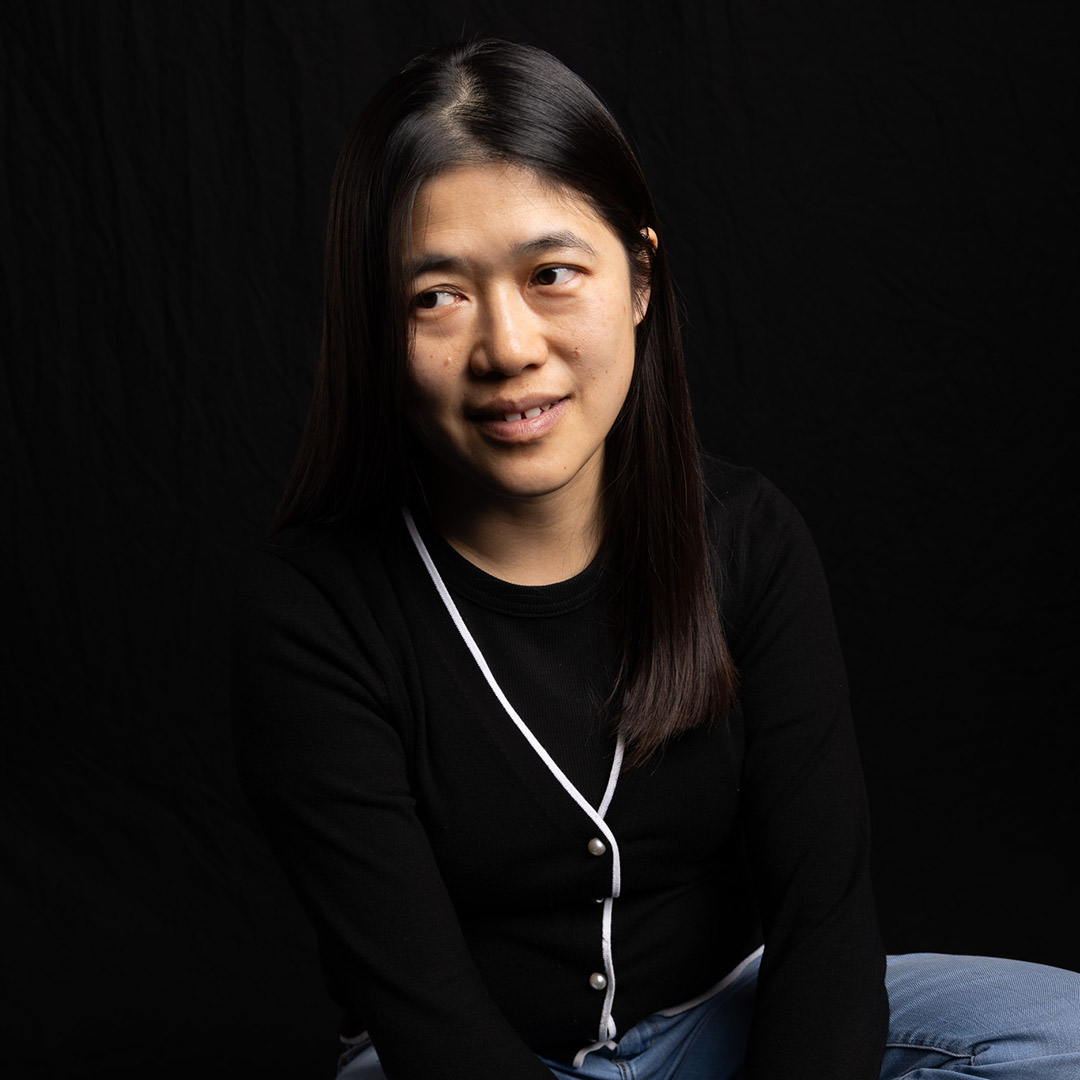 Jing is simling and looking off to the side. She has long dark hair and is wearing a black top with white trim and silver buttons with blue jeans. 