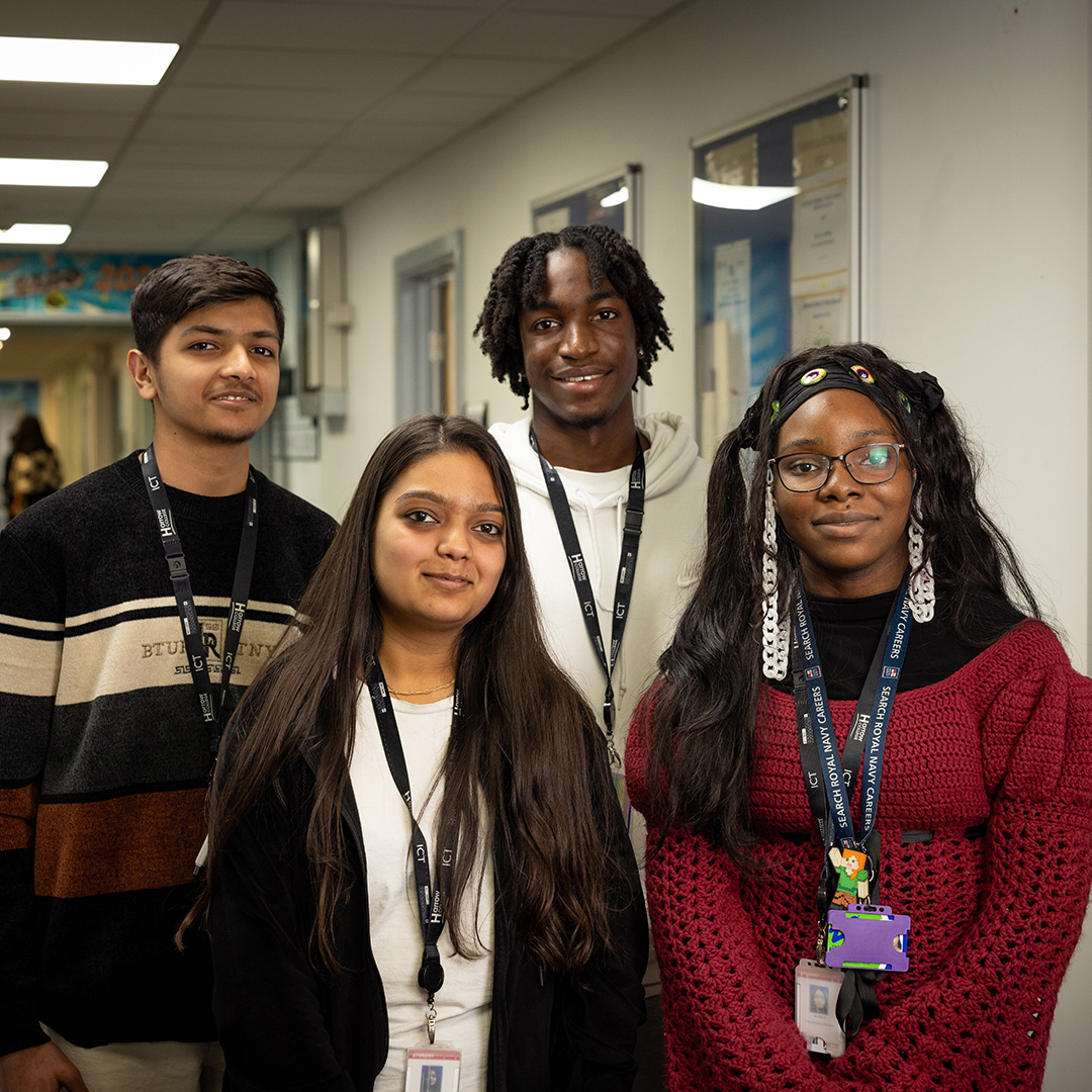 Four Harrow College students standing in the hall at Harrow College. They are all smiling and standing next to a paper image of a tree with photos of students. 