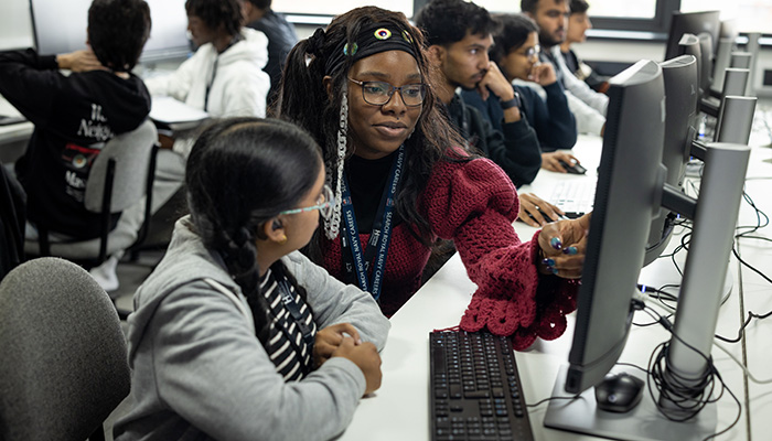Two female Harrow computing students working in the computing lab with other students around them. They are sitting in front of computer screens and keyboards.