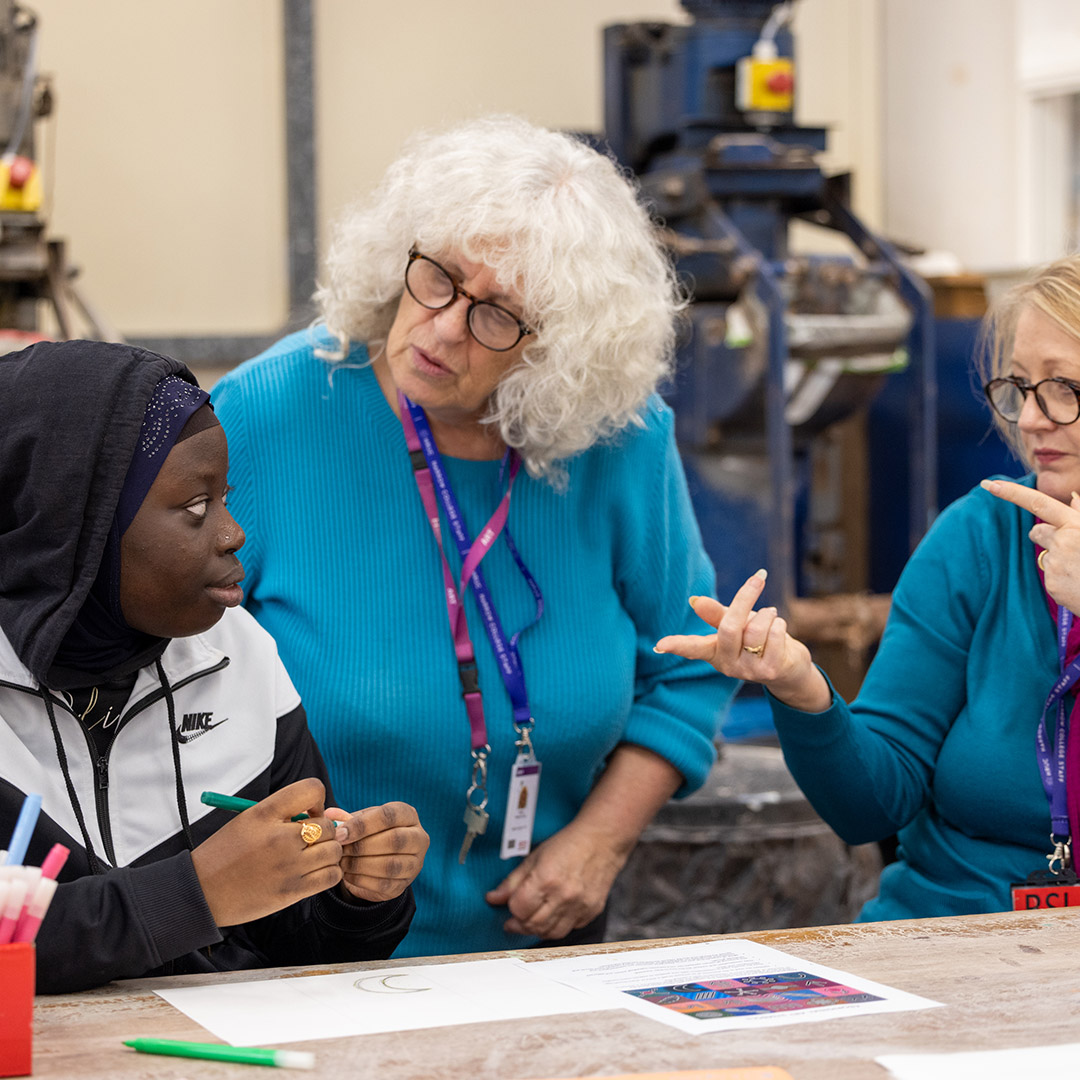 Harrow Weald SEN student working with two female staff members in a workshop on an art project. The staff members are both wearing blue tops and glasses.