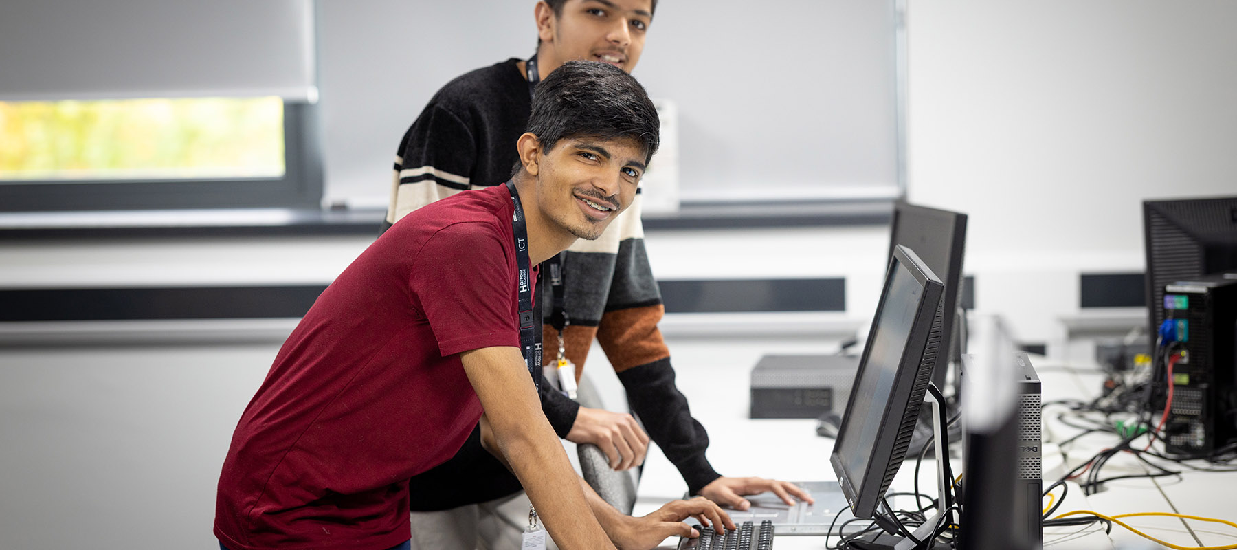 Two male Harrow College ICT students in front of a row of computers and smiling. The student standing in front is wearing a red t-shirt. 