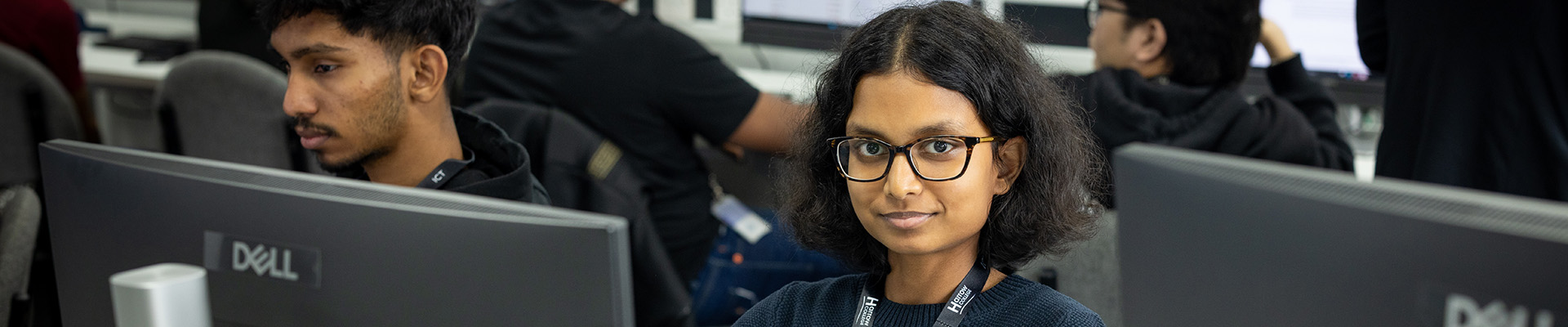 Female Harrow College student smiling. She is wearing glasses and sitting in front of a computer monitor in class with other students around her. 