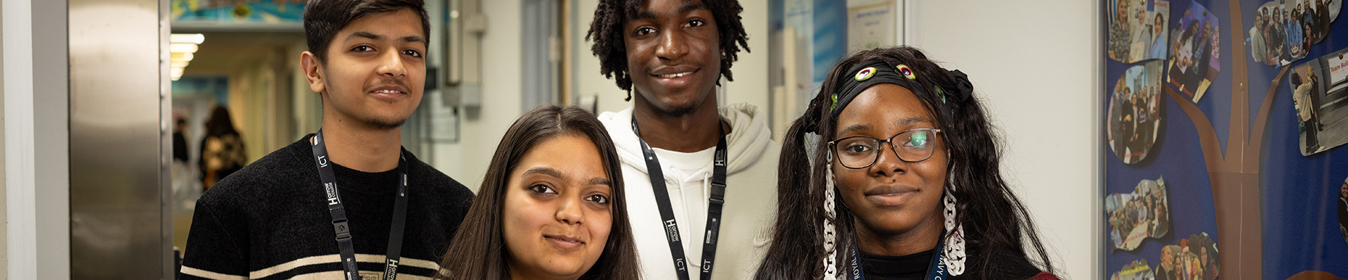 Four Harrow College students standing in the hall at Harrow College. They are all smiling and standing next to a paper image of a tree with photos of students. 