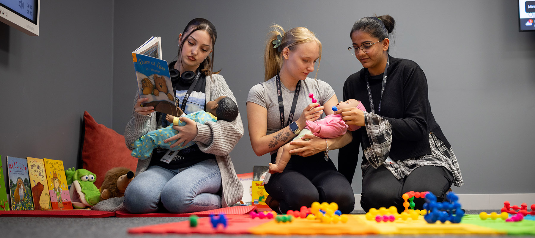 Three female Harrow College Health and Social Care students sitting on the ground with toys and children's books and practicing with baby dolls how to take care of them. 