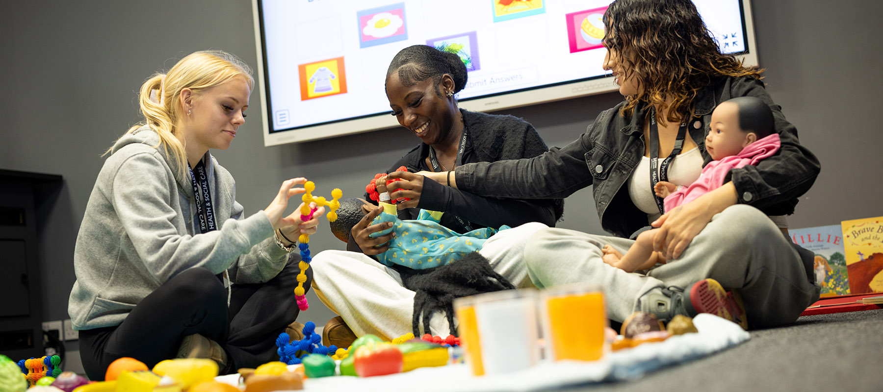 Harrow College Health and Social Care students sitting in a semi-circle on the floor and working with toys and baby dolls. There is a large screen behind them. 