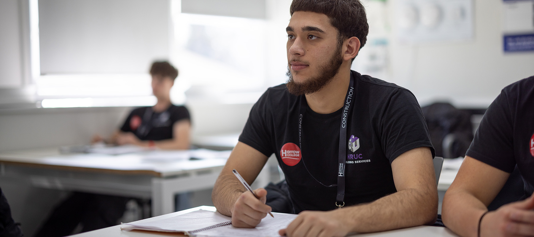 A male Harrow College Construction student sitting at a desk with a pen in his right hand ready to write in a notebook. He is in a classroom and looking ahead to the front of the class. He is wearing a black t-shirt with college logos on it. 