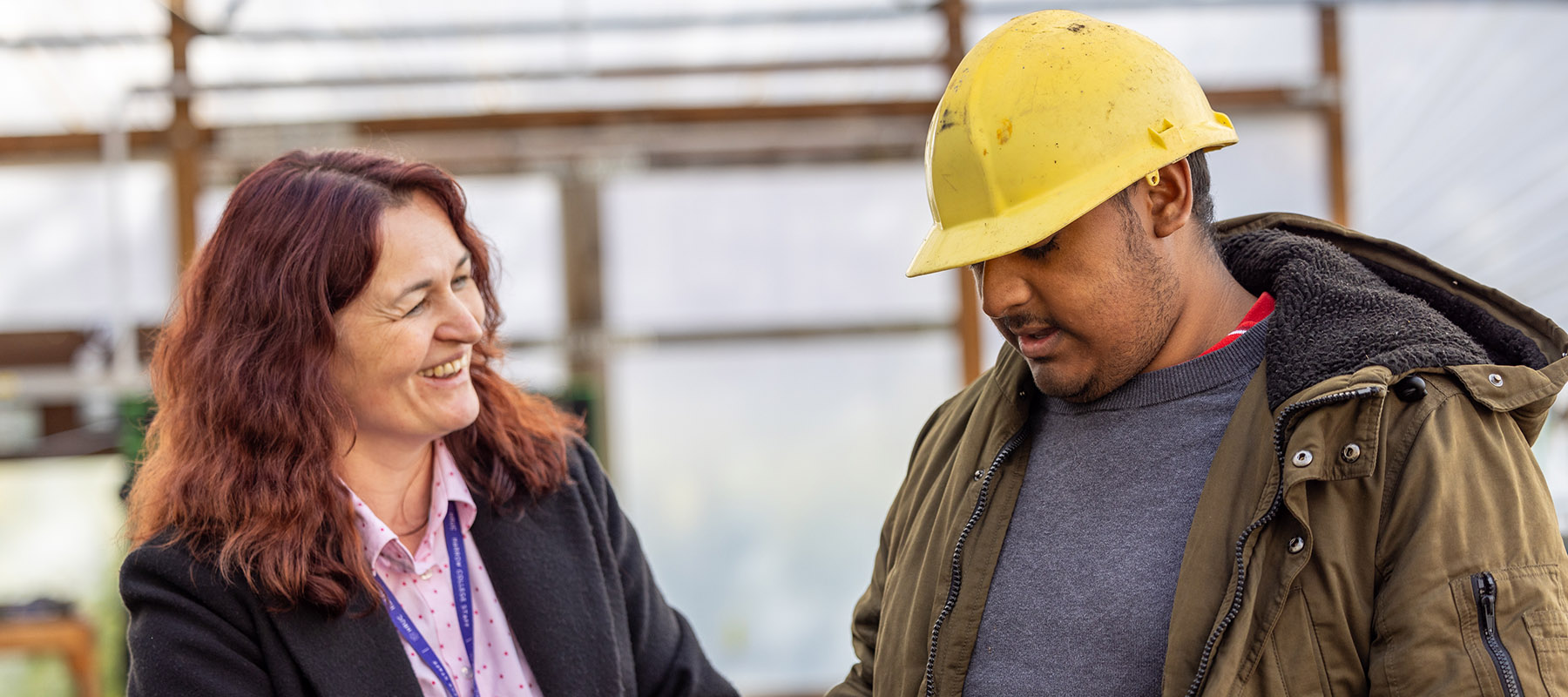 Harrow College student with a teacher at Harrow Weald campus. The male student is wearing a yellow hard hat and khaki jacket.