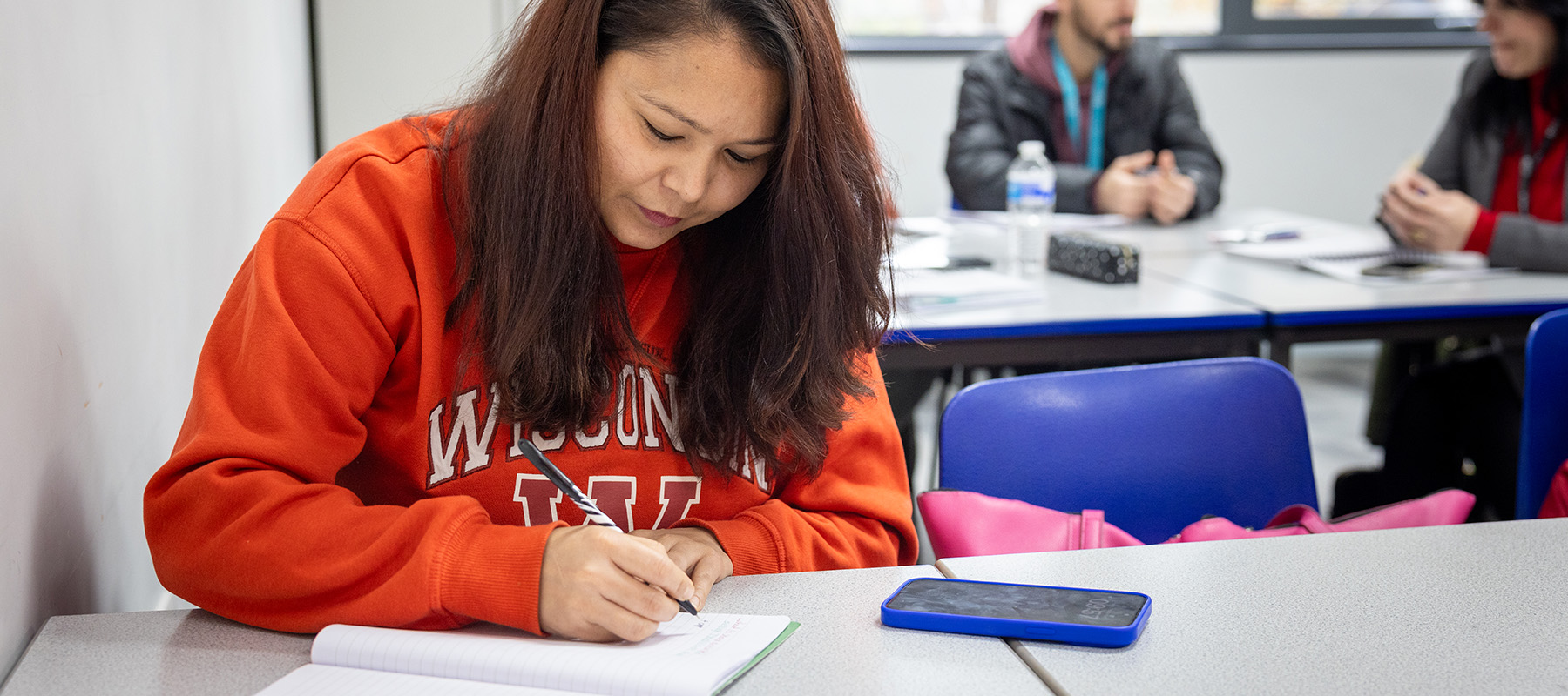 Female Harrow College student writing in a notepad. She is wearing an orange jumper and has a blue phone in front of her. There are students sitting behind her. 