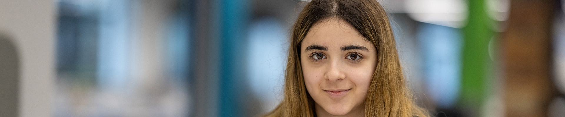 A female Harrow College Student smiling and sitting in the canteen. She has long brown hair.