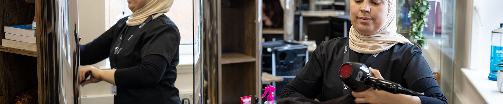 Female Harrow College Hairdressing student blow drying a woman's hair as she sits in front of a mirror in the Image Salon.