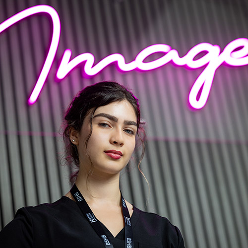 Female Harrow College Beauty student standing in front of the pink neon Image sign - she is wearing black with a black lanyard. 