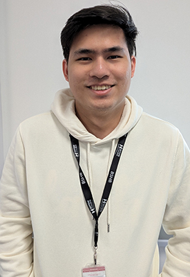 Luis Josef - International Student - smiling at the camera in front of a plain wall, wearing a white hoodie and black lanyard.