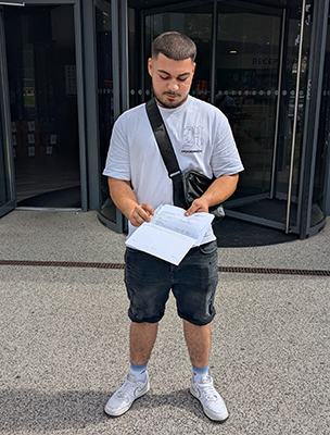 A male Harrow College student holding out his results letter and looking at it. He is standing outside the Harrow on the Hill College and wearing a white t-shirt with black shorts.