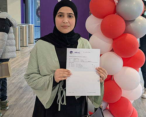 Female Harrow College student holding up her GCSE results letter in front of a welcome sign on a purple pillar and red and white balloons.