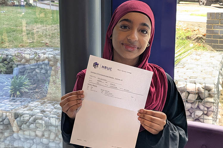 Female Harrow College student holding up her GCSE results letter in front of a purple pillar with windows behind her.