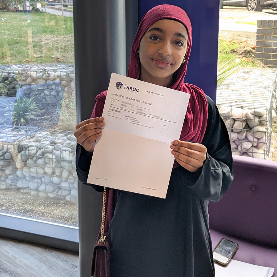 Female Harrow College student holding up her GCSE results letter in front of a purple pillar with windows behind her.