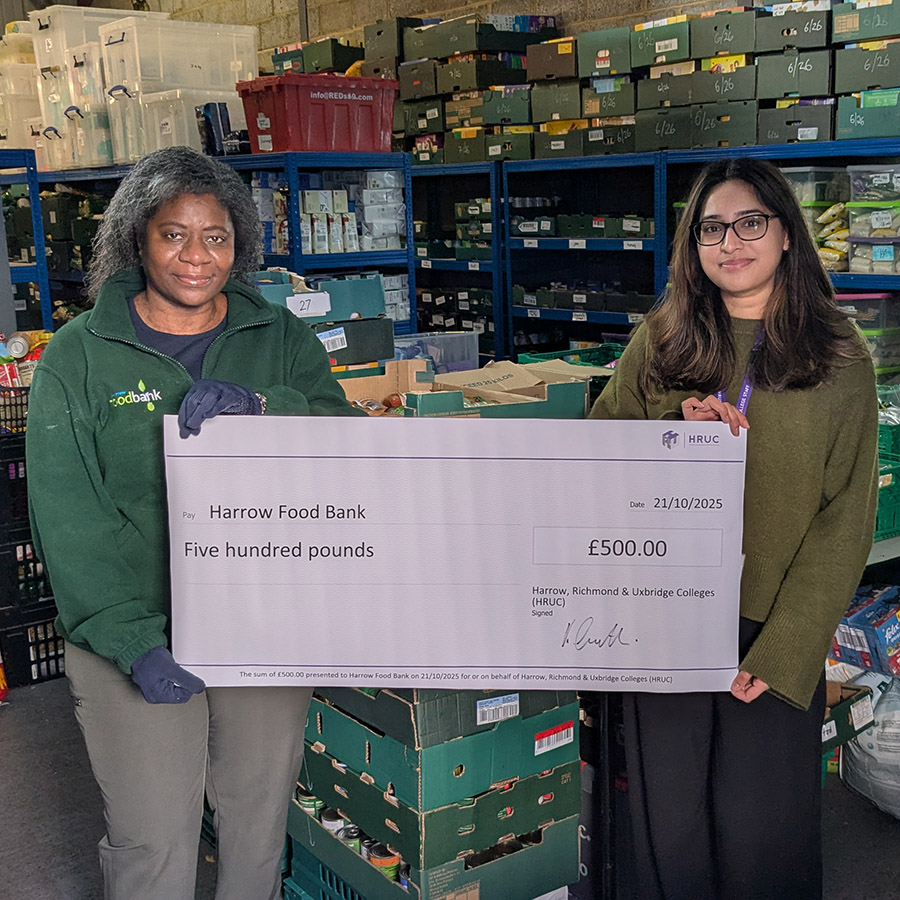 Yashvi Prajapati (Employee Experience Administrator)  presented a ceremonial cheque signed by HRUC CEO, Keith Smith, to Chonde Nkowani, Manager at Harrow Food Bank. They are standing in front of the shelves of food donations.