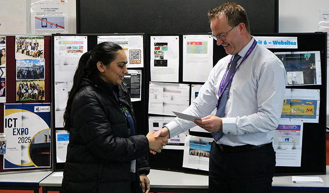 Chris Toms, Head of School, shaking the hand of a Harrow female student and giving her an award. They are standing in front of an ICT Expo display.