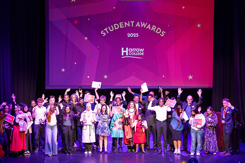 2025 Harrow College Student Awards evening - all award winners on stage, smiling and holding their hands and awards in the air. Standing in front of a big digital screen of a red awards sign.