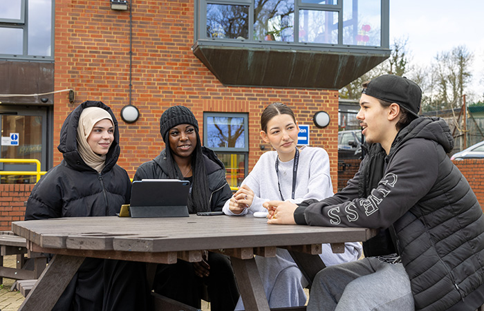 A group of students sitting outside at the Harrow Weald campus. They are sitting around a table talking and wearing coats and beanies. There is a brick building in the background. 