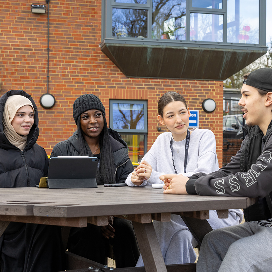 A group of students sitting outside at the Harrow Weald campus. They are sitting around a table talking and wearing coats and beanies. There is a brick building in the background. 