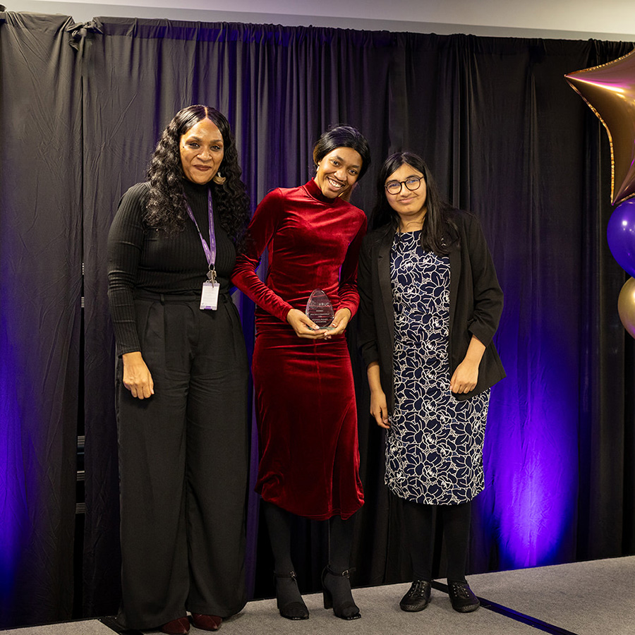 Business students from Harrow College winning an award at the HRUC Skills Competition Awards Ceremony. Presented by Kelly Ocloo, Assistant Principal. They are standing on stage in a group and smiling with one female student, in a red dress, holding the award. 
