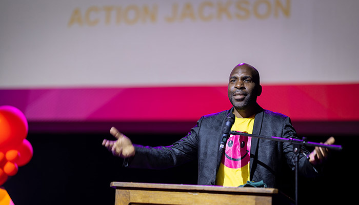 Action Jackson speaking on stage at the Harrow College Student Awards. His arms are outstretched and he is standing behind a lectern. There is a screen with his name behind him.