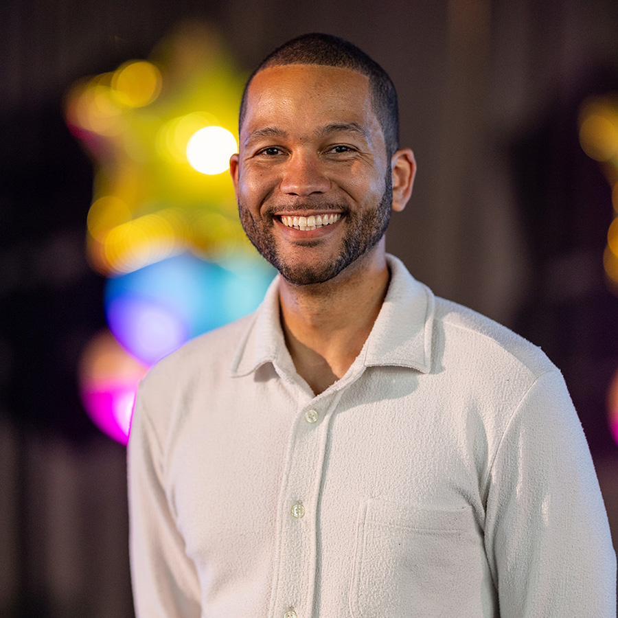 Anthony is smiling - he is wearing a white shirt and has close cropped hair and a beard. There are bright balloons behind him.