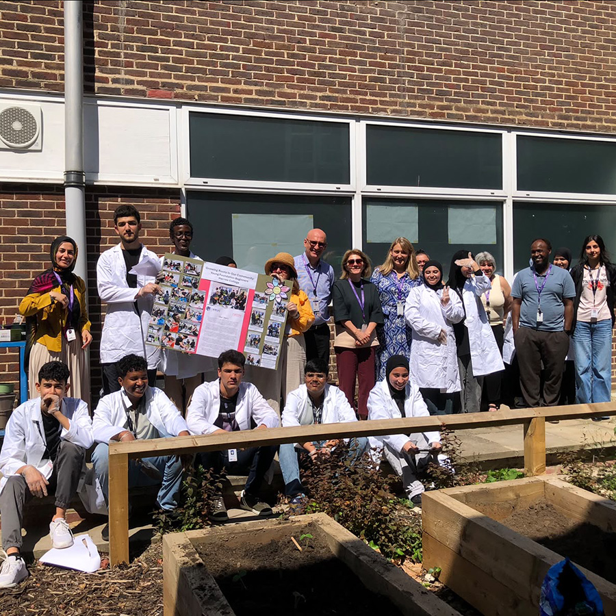 Harrow College ESOL students with teaching staff gathered in front of the Northwood Hills Underground community garden project. Some are wearing white lab coats to over their clothes. 