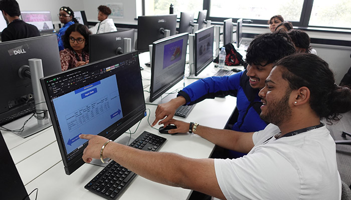Male Harrow College ICT students participating in the ICT Expo. They are sitting in a computer lab classroom with other students at a bank of computers and pointing at the screen.