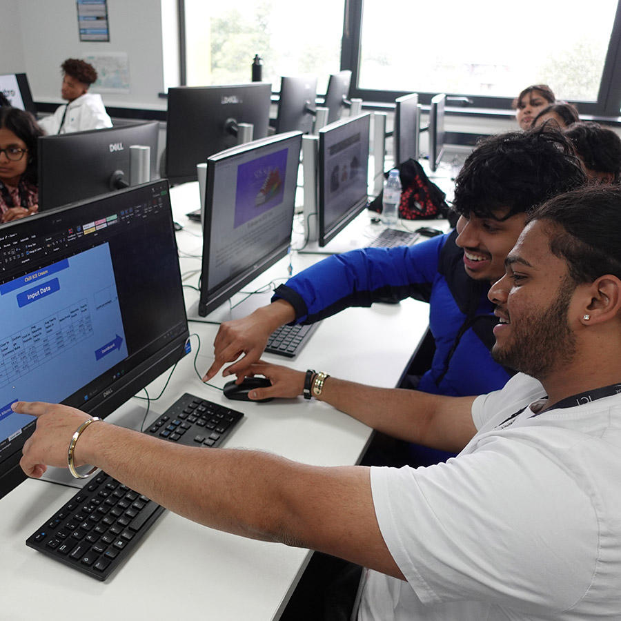 Male Harrow College ICT students participating in the ICT Expo. They are sitting in a computer lab classroom with other students at a bank of computers and pointing at the screen.