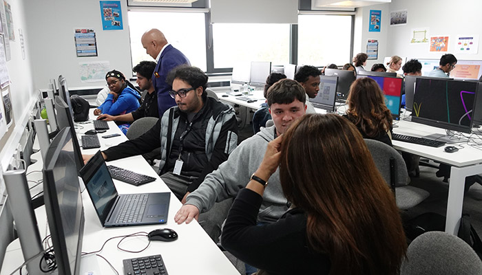 Students participating in the Harrow College ICT Expo. They are sitting in a computer lab working on projects. Teachers are looking at their progress or talking to the students.