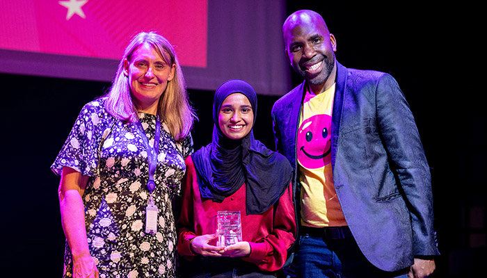 Our Harrow College Student Awards winner for the Greatest Contribution to College Life -Sidrah - standing with Jo Swindells, Principal of Harrow College and Action Jackson, who was a guest speaker at the awards. Sidrah is holding her award and standing between Jo and Action Jackson with a red screen background behind them. 