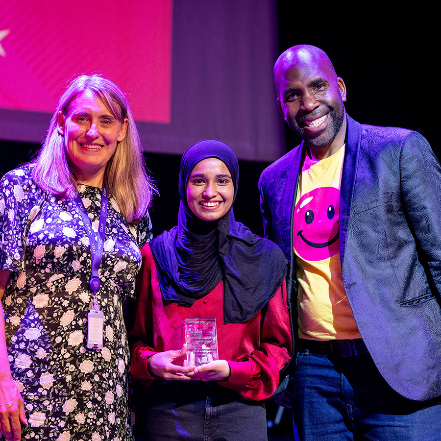 Our Harrow College Student Awards winner for the Greatest Contribution to College Life -Sidrah - standing with Jo Swindells, Principal of Harrow College and Action Jackson, who was a guest speaker at the awards. Sidrah is holding her award and standing between Jo and Action Jackson with a red screen background behind them. 
