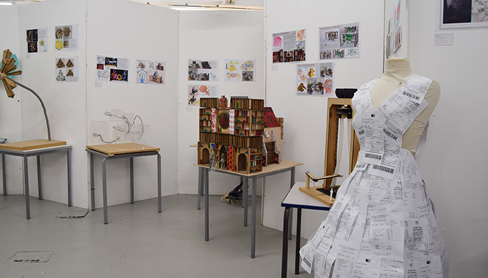 Harrow College Art and Design students showcasing their portfolio of work with models, designs and art pieces in an end-of-year showcase. There are designs displayed on walls and models on tables with a dressmaker mannequin using receipts as a dress in the foreground. 