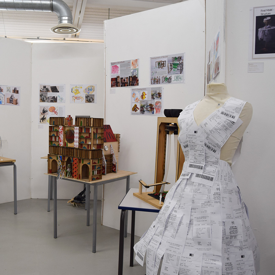 Harrow College Art and Design students showcasing their portfolio of work with models, designs and art pieces in an end-of-year showcase. There are designs displayed on walls and models on tables with a dressmaker mannequin using receipts as a dress in the foreground. 