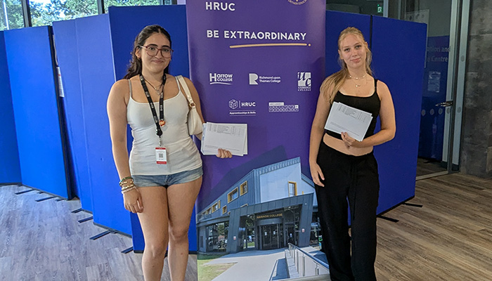 Two female Harrow College students holding their exam results and standing either side of the Be Extraordinary purple pull-up banner in reception at Harrow on the Hill campus.