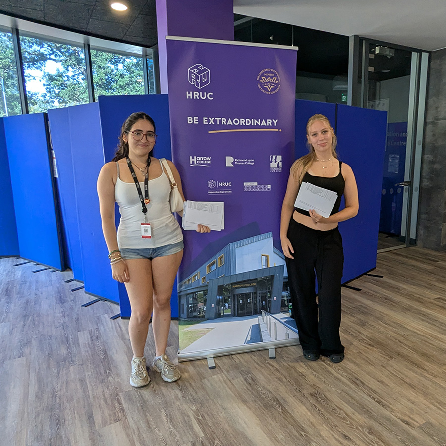 Two female Harrow College students holding their exam results and standing either side of the Be Extraordinary purple pull-up banner in reception at Harrow on the Hill campus.