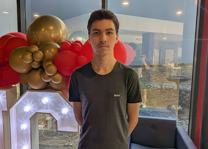 Male Harrow College student standing in Harrow on the Hill College reception after receving his GCSE exam results. There is a lit up sign behind him with the letter C visible and red and gold balloons on top of the sign. He is wearing a grey t-shirt and blue track suit pants. 
