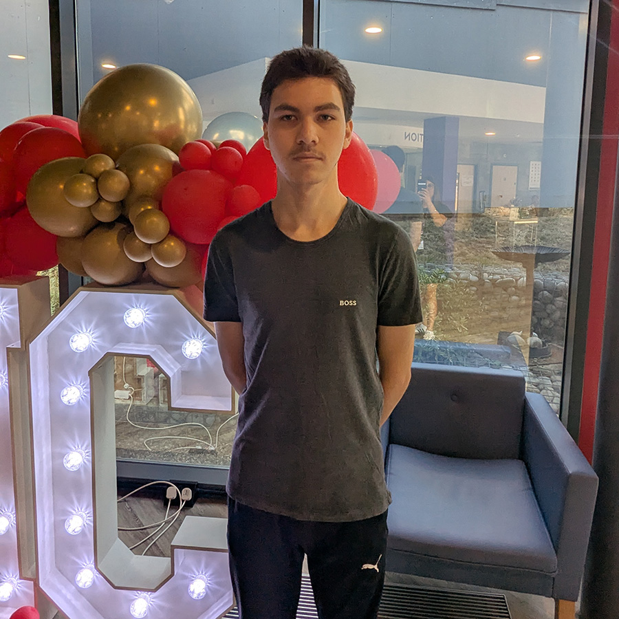 Male Harrow College student standing in Harrow on the Hill College reception after receving his GCSE exam results. There is a lit up sign behind him with the letter C visible and red and gold balloons on top of the sign. He is wearing a grey t-shirt and blue track suit pants. 