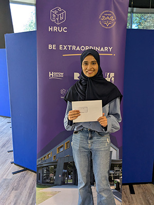 Harrow College student Sidrah is holding her A-Levels results envelope and standing in Harrow College reception in front of a Be Extraordinary purple banner. She is smiling.