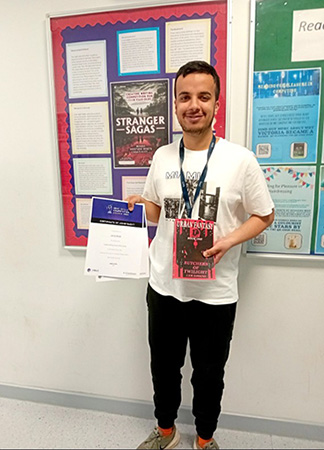 Jamal Khan - prize winner of the Young Writers Stranger Sagas competition. Holding his certificate and a book in front of a noticeboard with the Stranger Sagas poster.