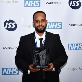 Idris Mohammed, pictured, holding his award, dressed in a black suit with an NHS graphic background